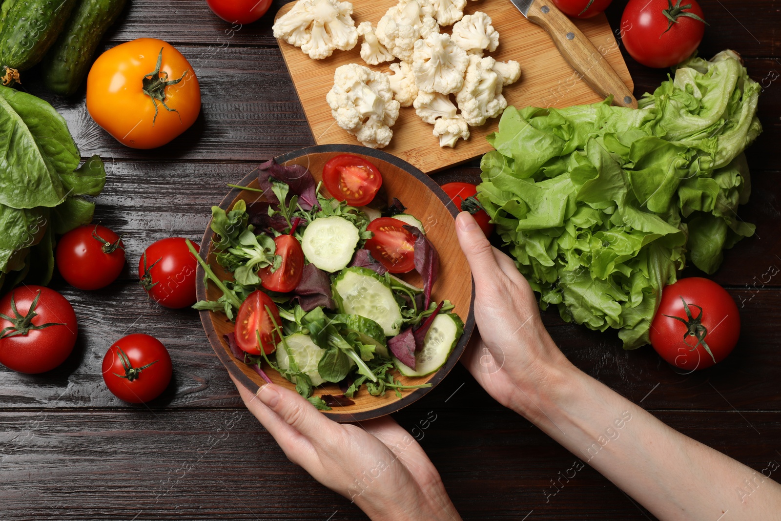 Photo of Healthy vegetarian food. Woman holding bowl of salad at wooden table with vegetables, top view