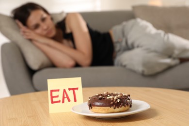 Photo of Eating disorder. Woman lying on sofa indoors, focus on sticky note with word Eat and donut