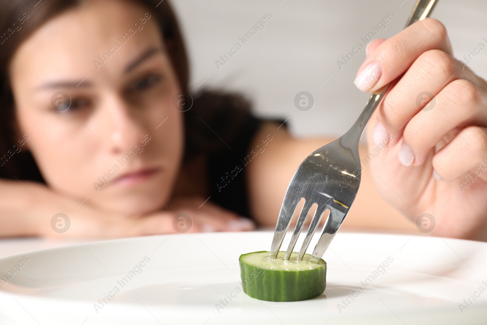 Photo of Eating disorder. Sad woman holding fork with cucumber at table indoors, selective focus
