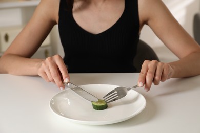 Photo of Eating disorder. Woman cutting cucumber at white table indoors, closeup