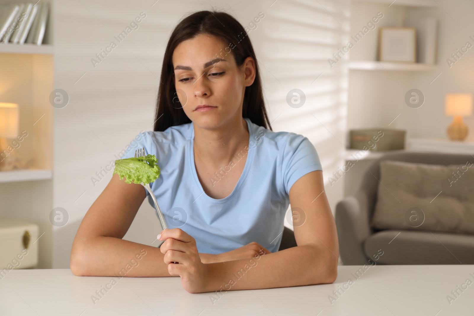 Photo of Eating disorder. Sad woman holding fork with lettuce at white table indoors