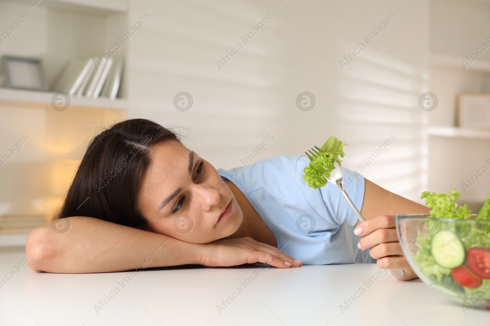 Photo of Eating disorder. Sad woman holding fork with lettuce at white table indoors