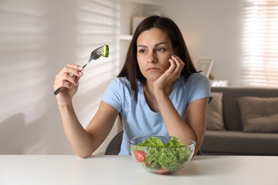 Eating disorder. Sad woman holding fork with cucumber at white table indoors