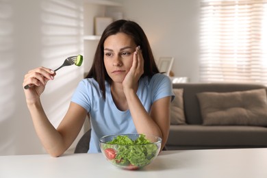 Photo of Eating disorder. Sad woman holding fork with cucumber at white table indoors