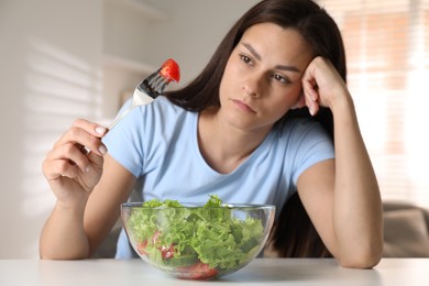 Photo of Eating disorder. Sad woman holding fork with tomato over bowl at white table indoors