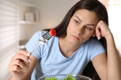 Eating disorder. Sad woman holding fork with tomato indoors