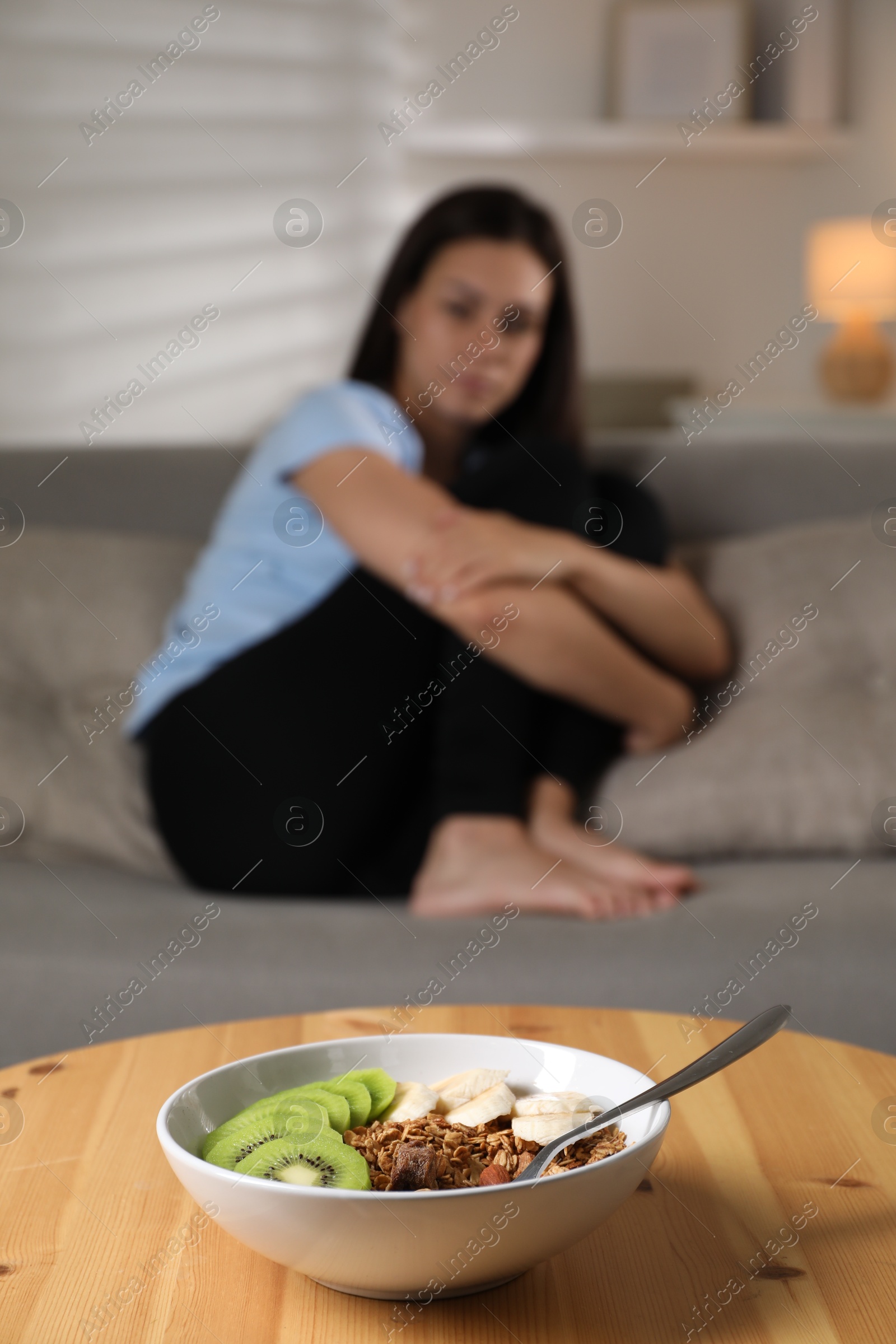 Photo of Eating disorder. Sad woman sitting on sofa indoors, focus on granola, kiwi and banana