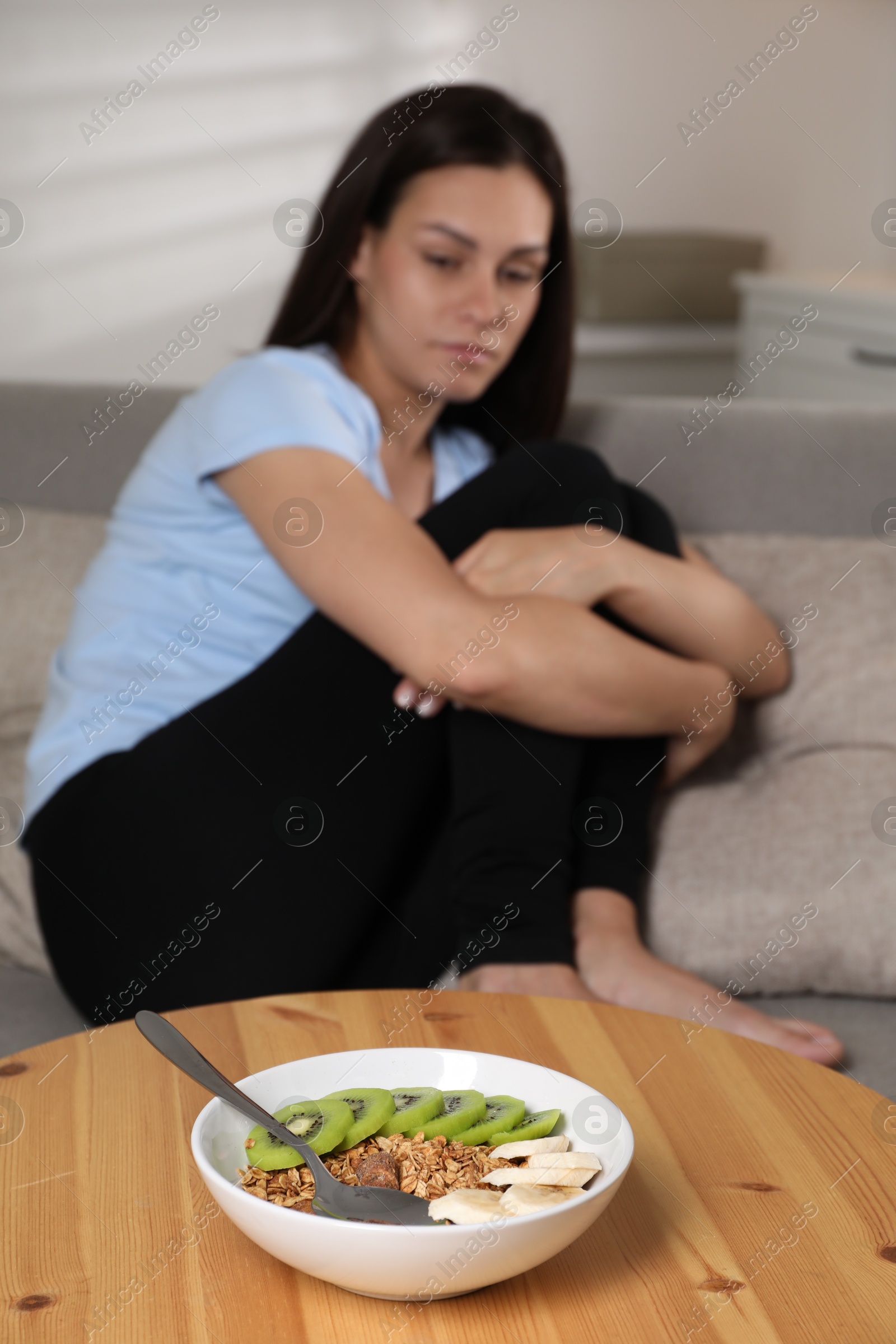 Photo of Eating disorder. Sad woman on sofa indoors, focus on granola, kiwi and banana