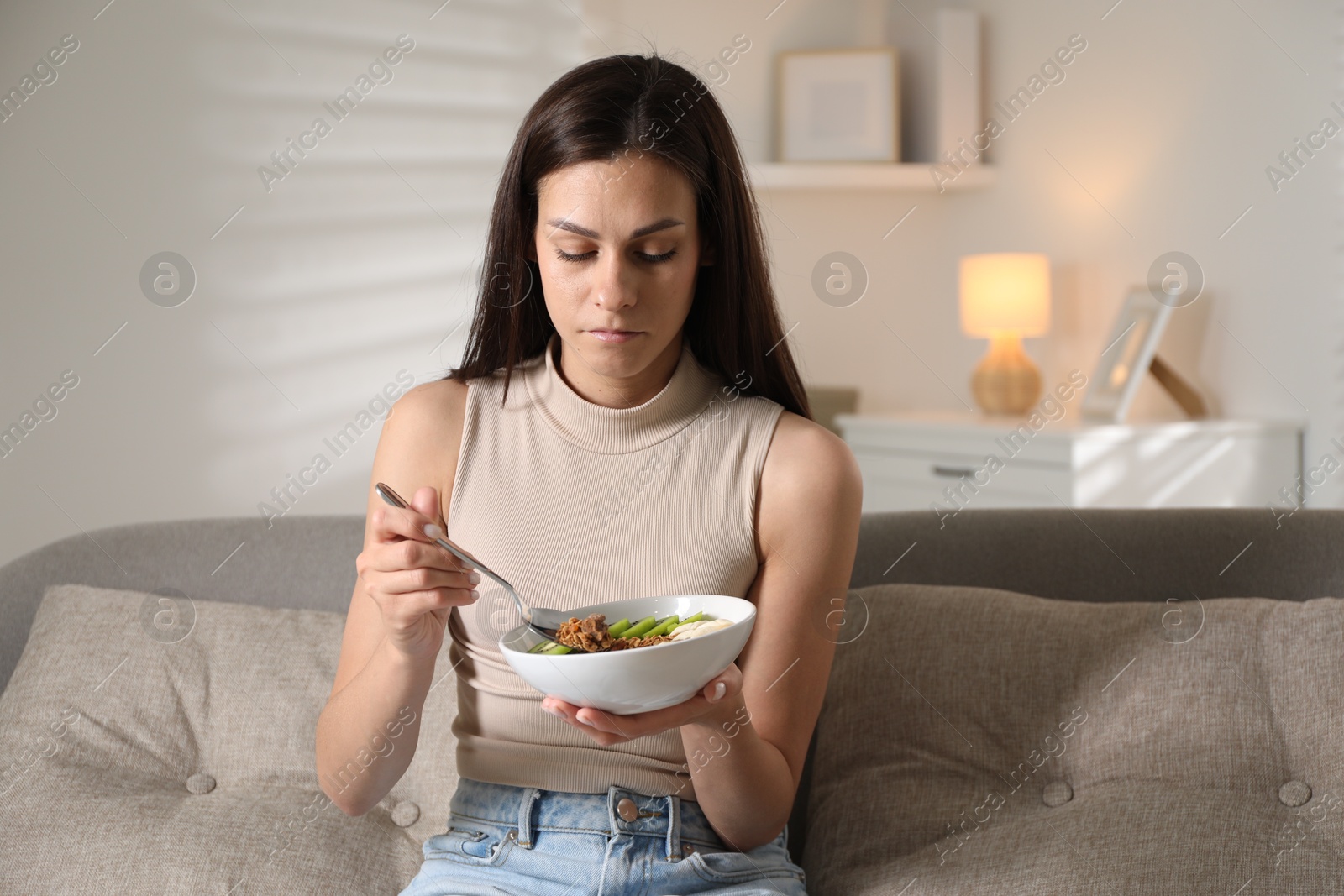 Photo of Eating disorder. Sad woman holding spoon with granola and bowl on sofa indoors