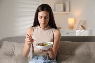 Photo of Eating disorder. Sad woman holding spoon with granola and bowl on sofa indoors