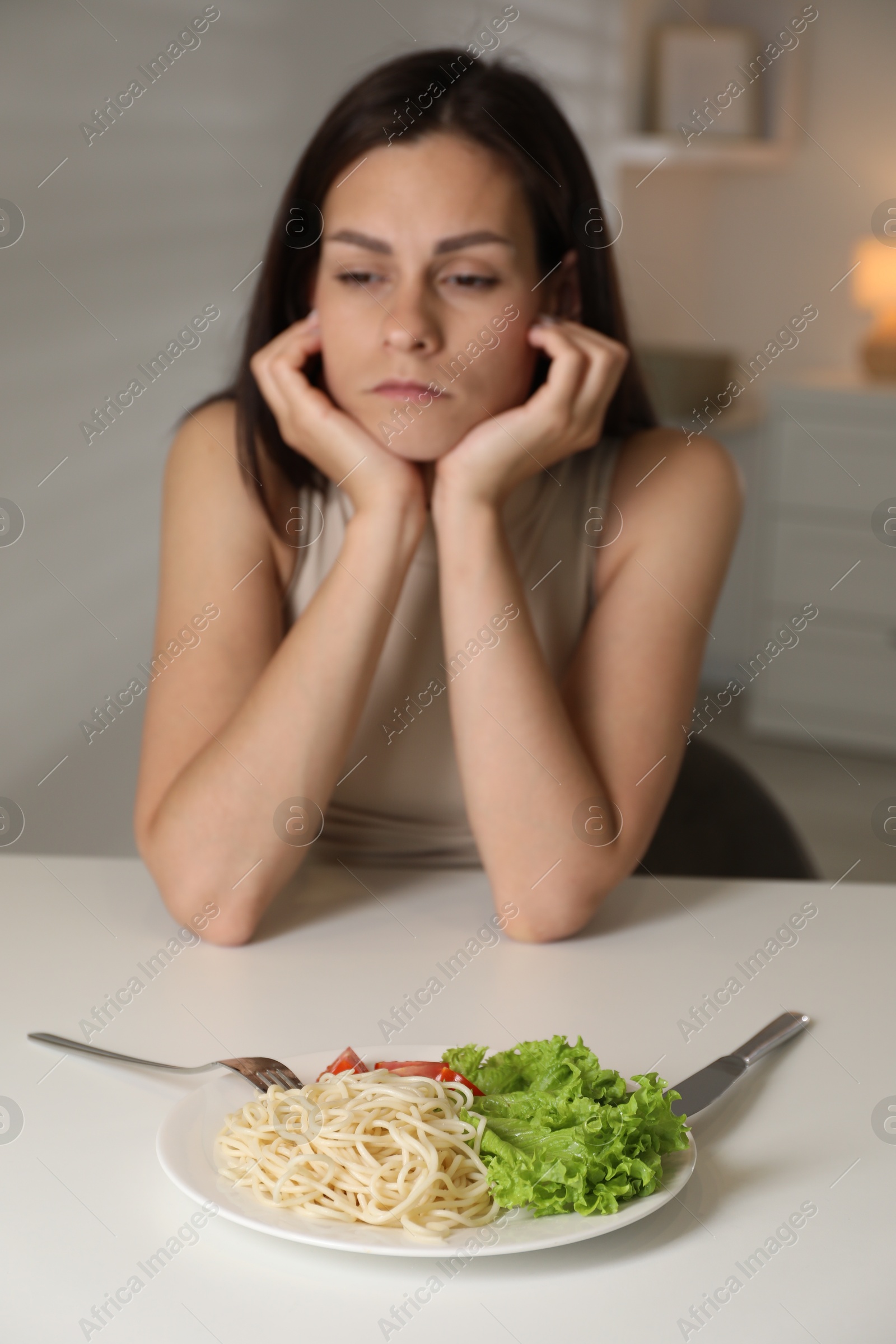 Photo of Eating disorder. Sad woman at white table with spaghetti and cutlery indoors