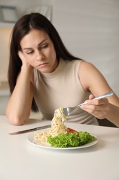 Eating disorder. Sad woman holding fork with spaghetti over plate at white table indoors