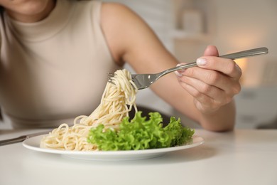 Eating disorder. Woman holding fork with spaghetti over plate at white table indoors, closeup