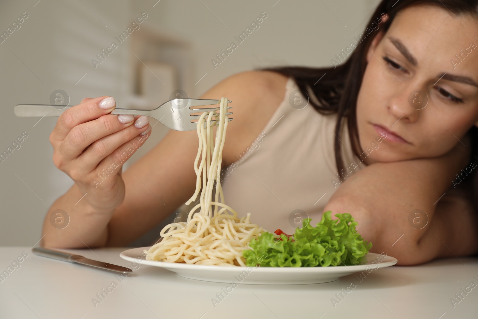 Photo of Eating disorder. Sad woman holding fork with spaghetti over plate at white table indoors