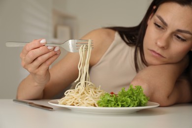 Eating disorder. Sad woman holding fork with spaghetti over plate at white table indoors