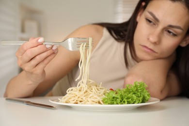 Eating disorder. Sad woman holding fork with spaghetti over plate at white table indoors