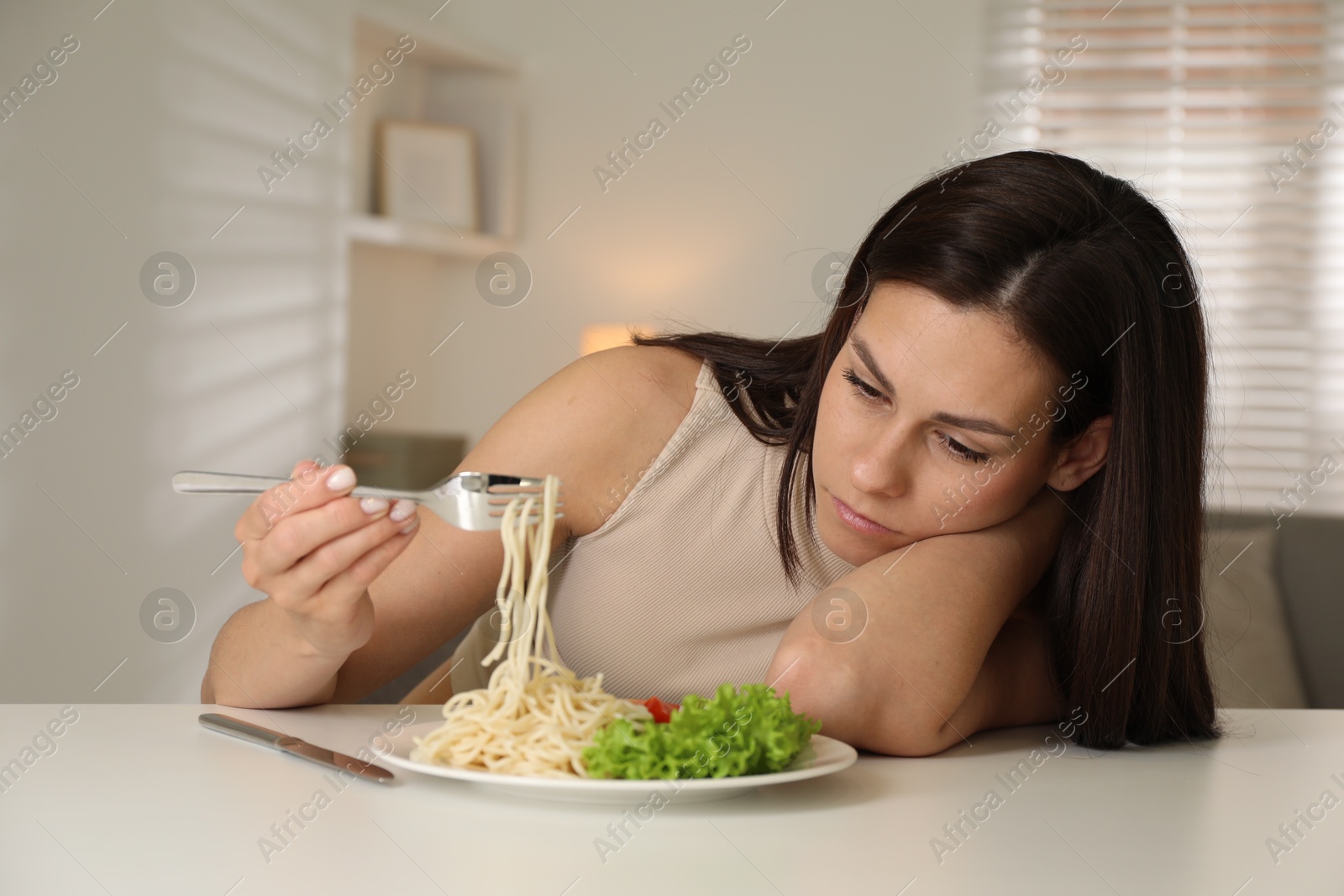 Photo of Eating disorder. Sad woman holding fork with spaghetti over plate at white table indoors
