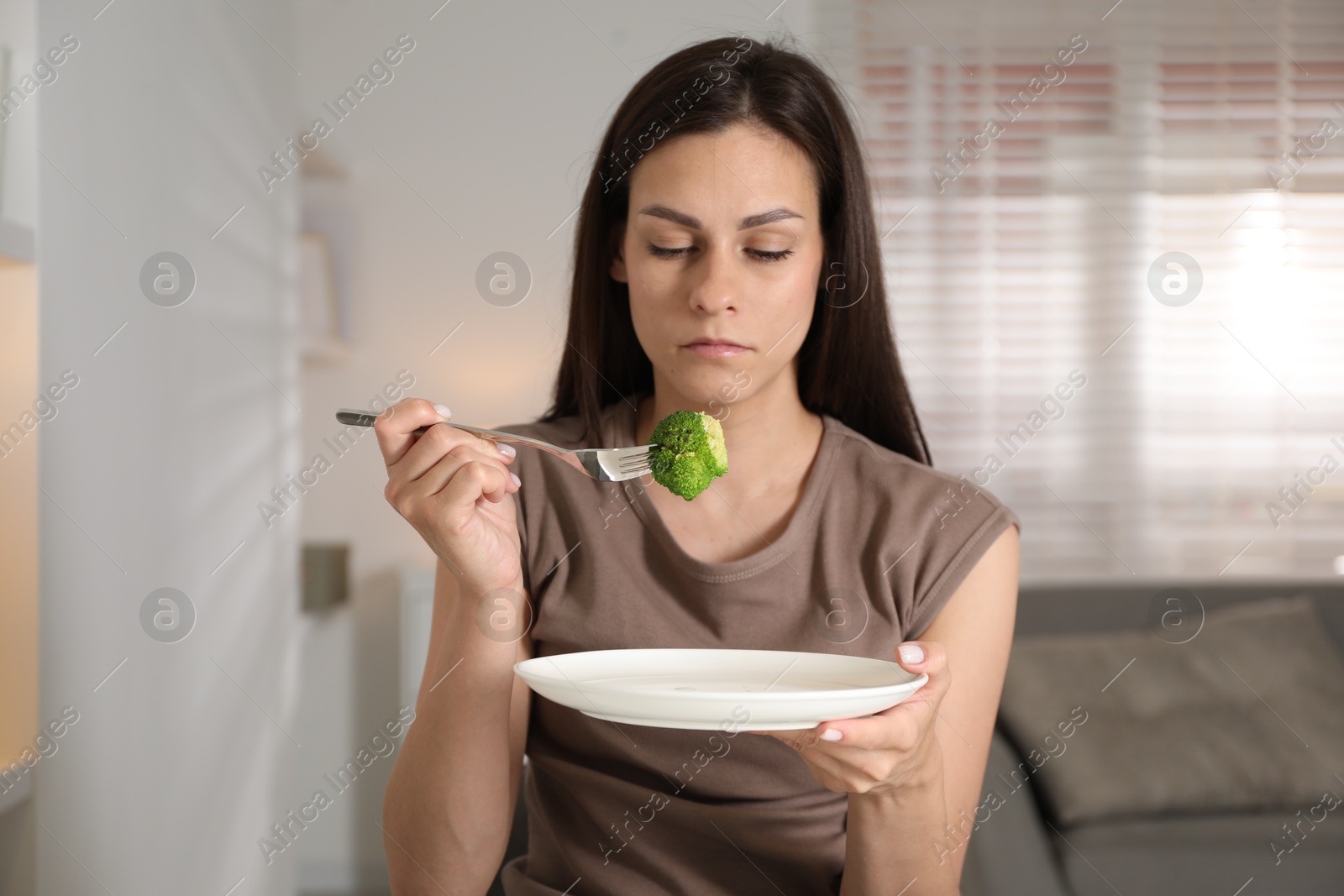 Photo of Eating disorder. Sad woman holding fork with broccoli over plate indoors