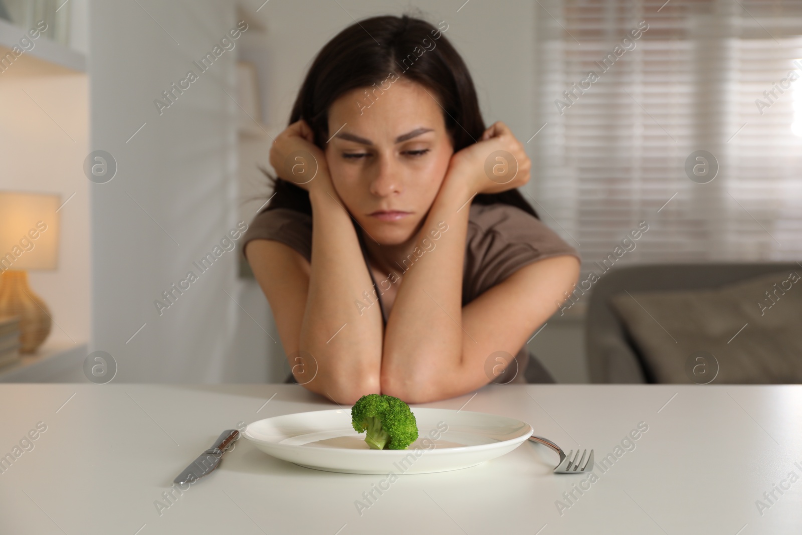 Photo of Eating disorder. Sad woman at white table with cutlery, broccoli and plate indoors