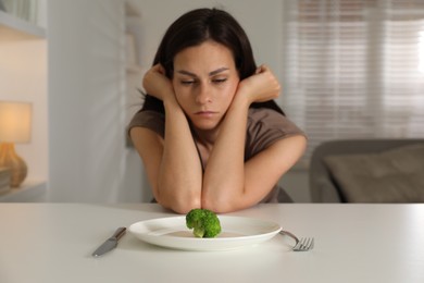 Eating disorder. Sad woman at white table with cutlery, broccoli and plate indoors