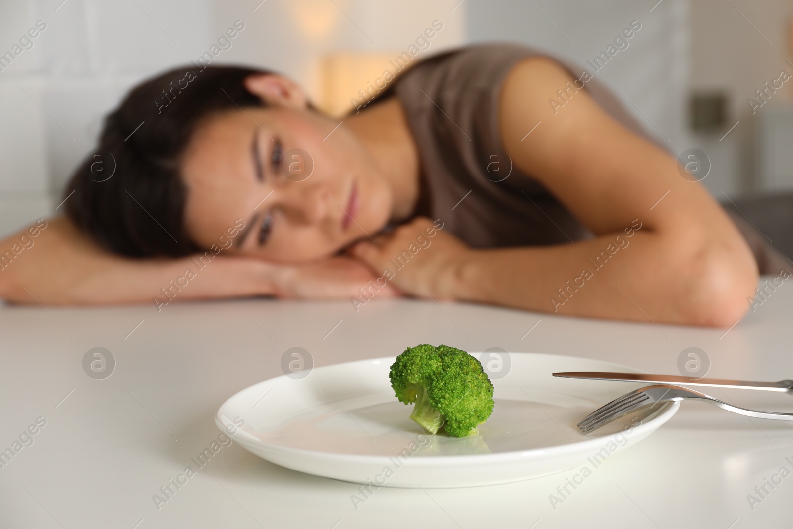 Photo of Eating disorder. Sad woman at white table, focus on broccoli and cutlery