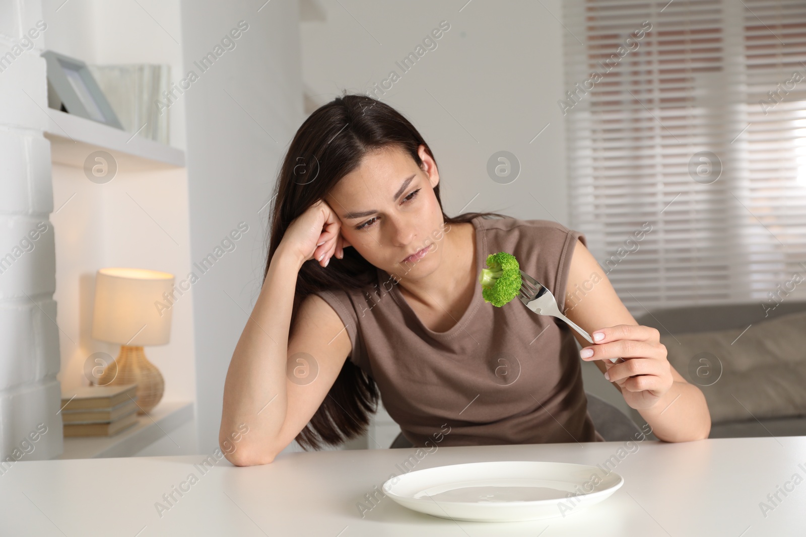 Photo of Eating disorder. Sad woman holding fork with broccoli at white table indoors