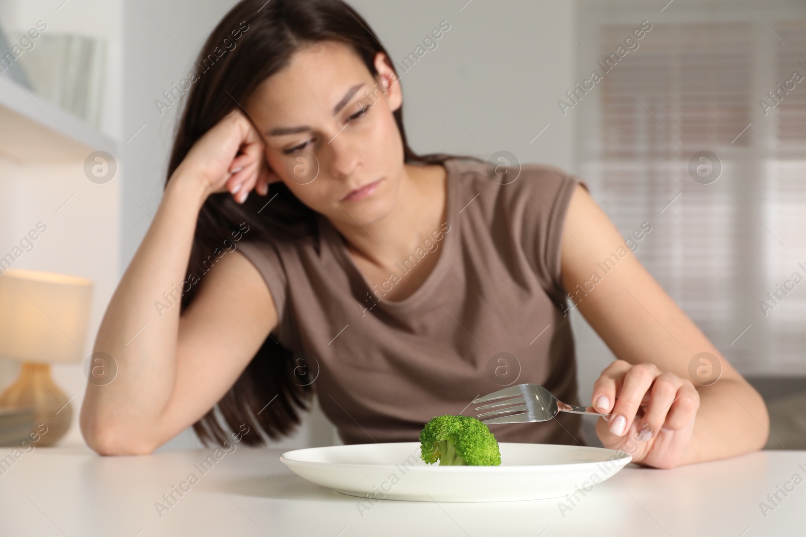 Photo of Eating disorder. Sad woman holding fork near broccoli at table indoors