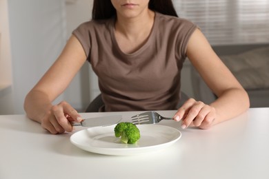 Photo of Eating disorder. Woman holding cutlery near broccoli at white table indoors, closeup