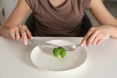 Eating disorder. Woman holding cutlery near broccoli at white table indoors, closeup