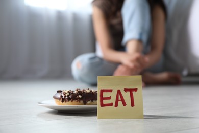 Photo of Eating disorder. Woman sitting on floor indoors, focus on sticky note with word Eat and donut