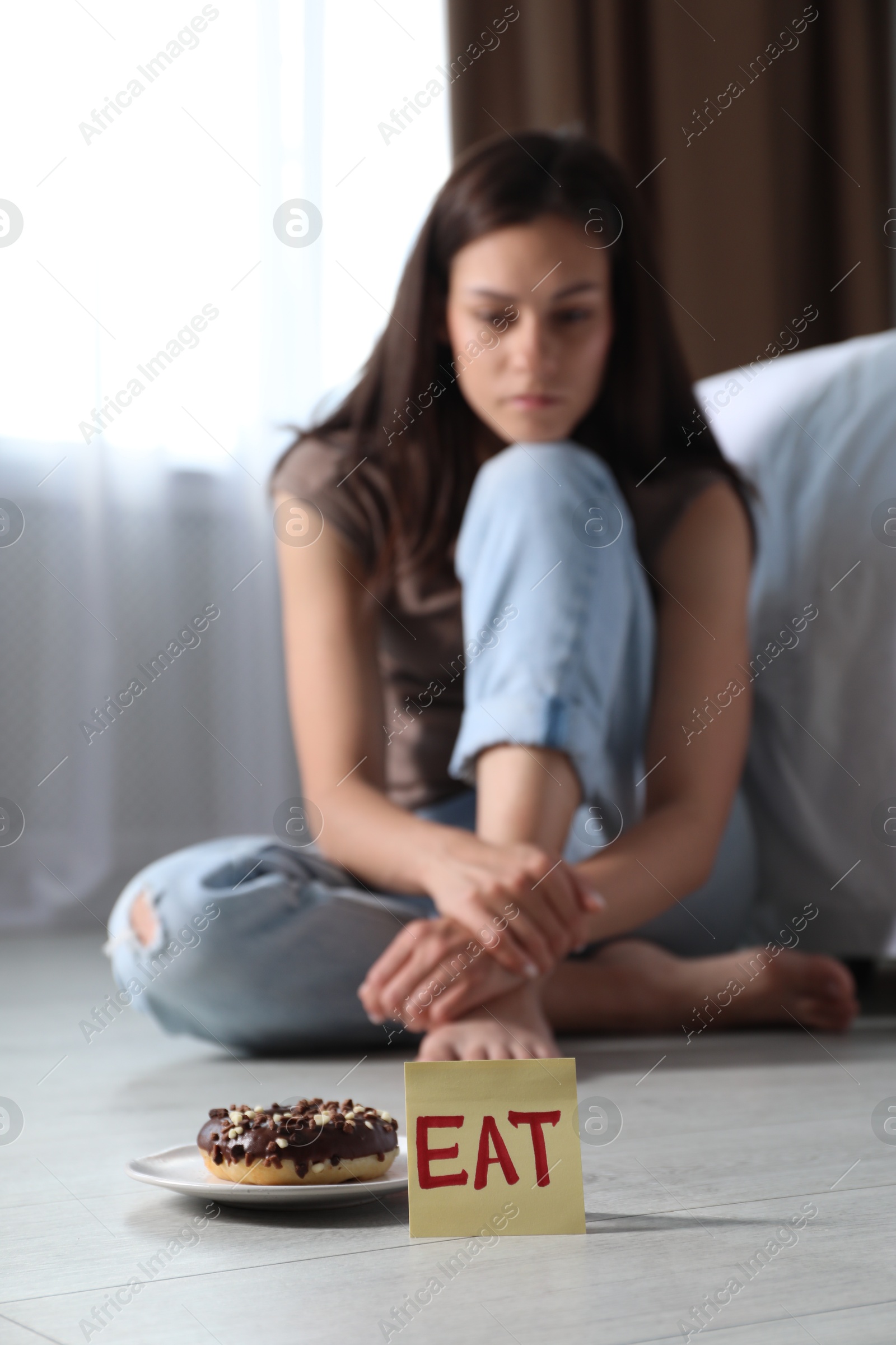 Photo of Eating disorder. Sad woman sitting on floor indoors, focus on note with word Eat and donut