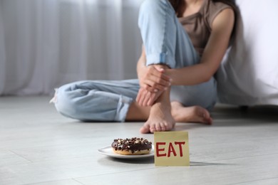 Photo of Eating disorder. Woman sitting on floor indoors, focus on sticky note with word Eat and donut