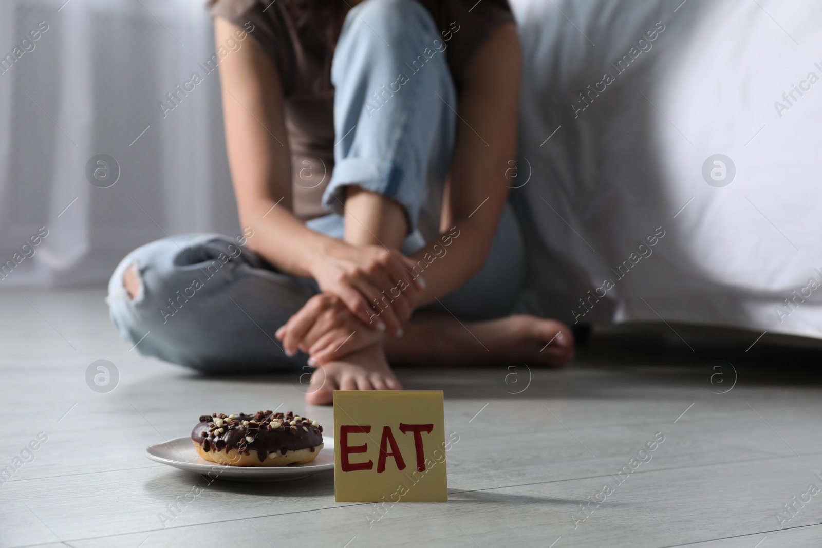Photo of Eating disorder. Woman sitting on floor indoors, focus on sticky note with word Eat and donut