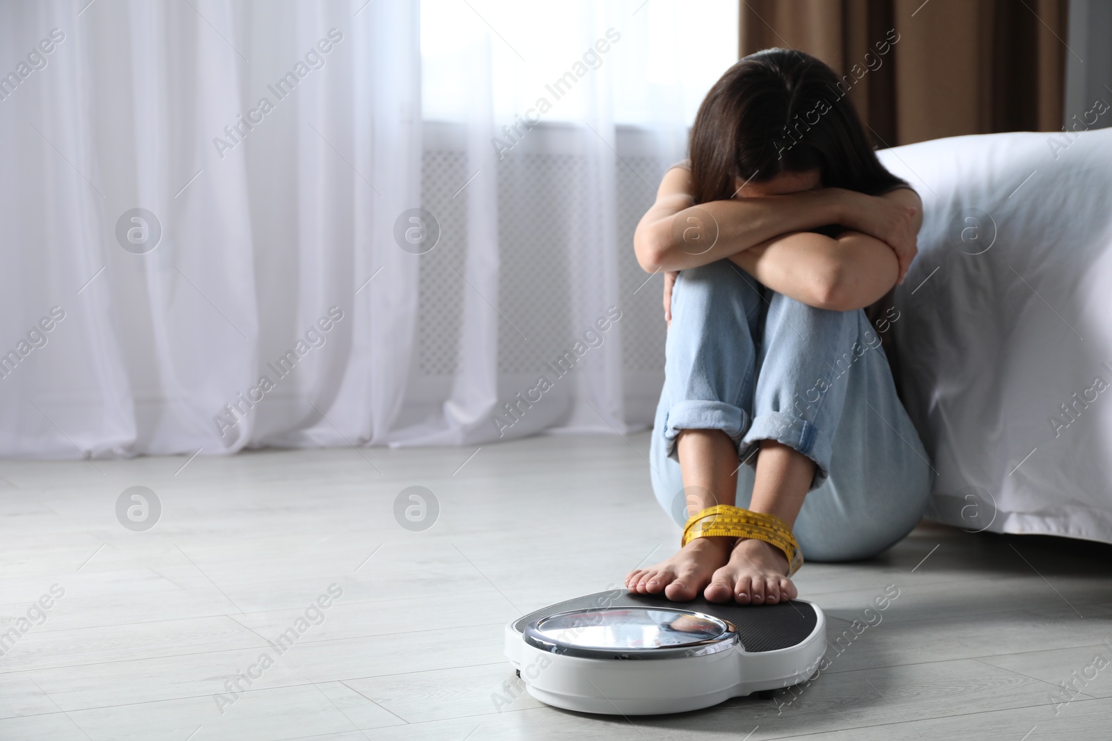 Photo of Eating disorder. Depressed woman sitting near scale on floor indoors, space for text