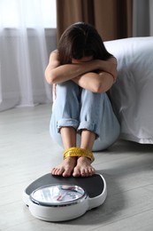 Photo of Eating disorder. Depressed woman sitting near scale on floor indoors