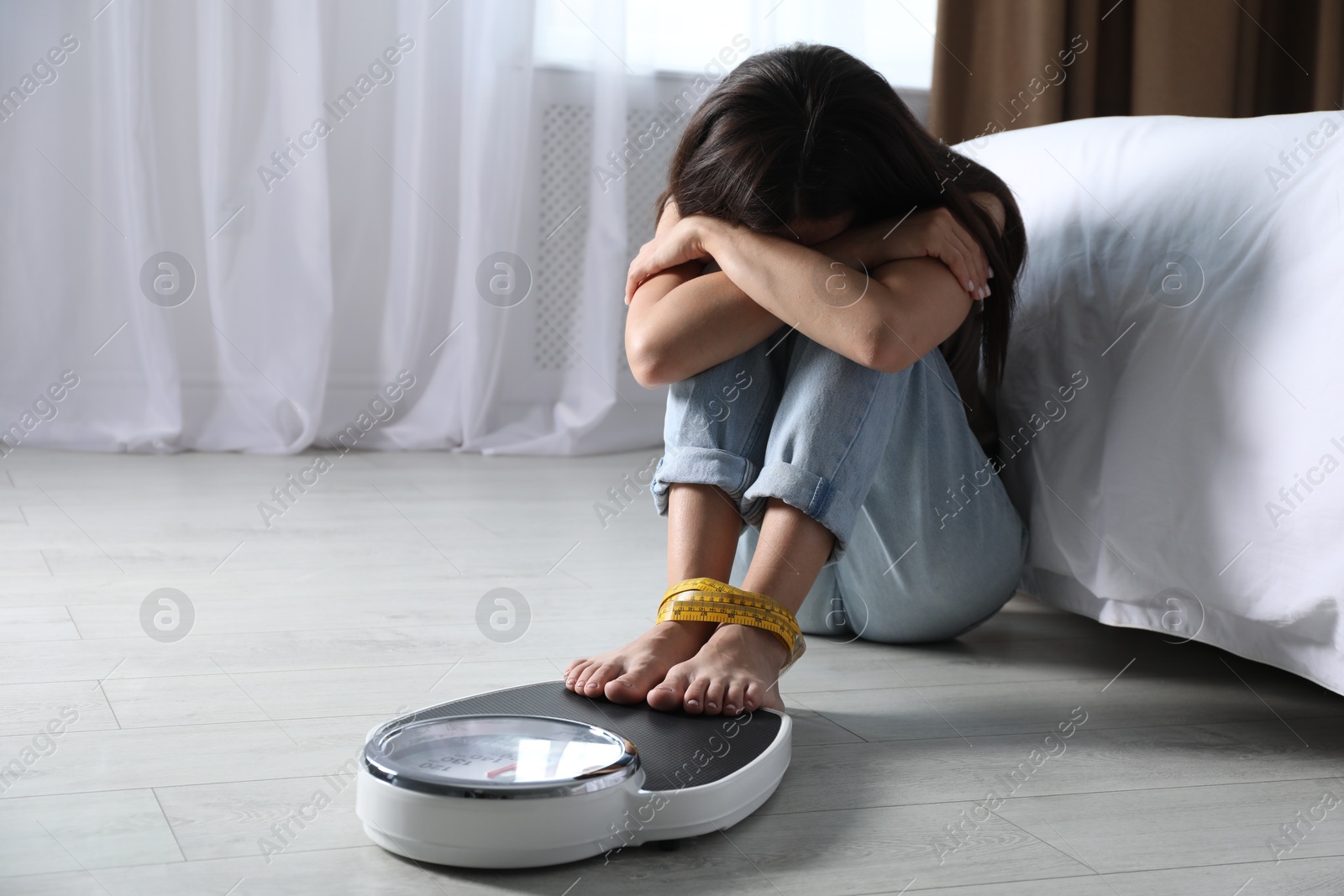 Photo of Eating disorder. Depressed woman sitting near scale on floor indoors, space for text