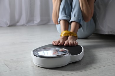 Eating disorder. Woman sitting near scale on floor indoors, closeup