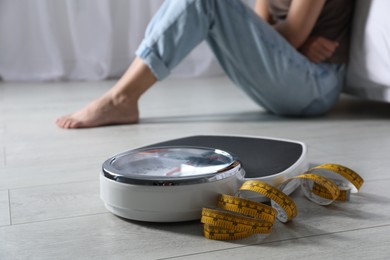 Eating disorder. Sad woman sitting near scale and measuring tape on floor indoors, selective focus