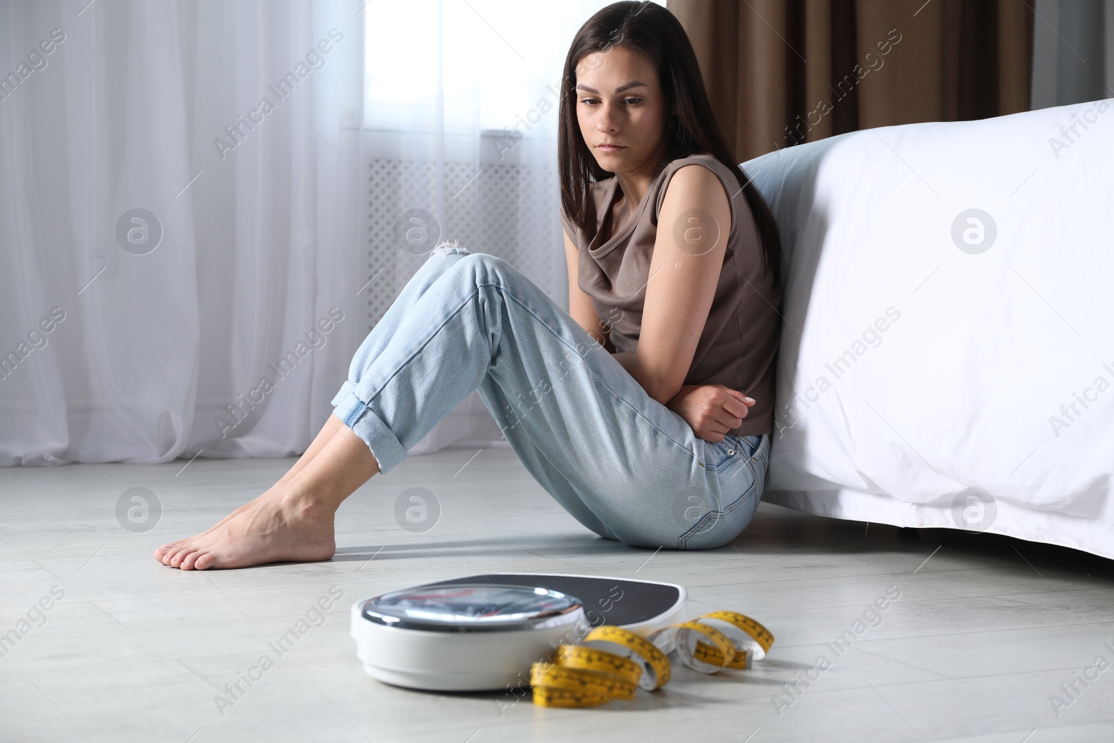 Photo of Eating disorder. Sad woman sitting near scale and measuring tape on floor indoors