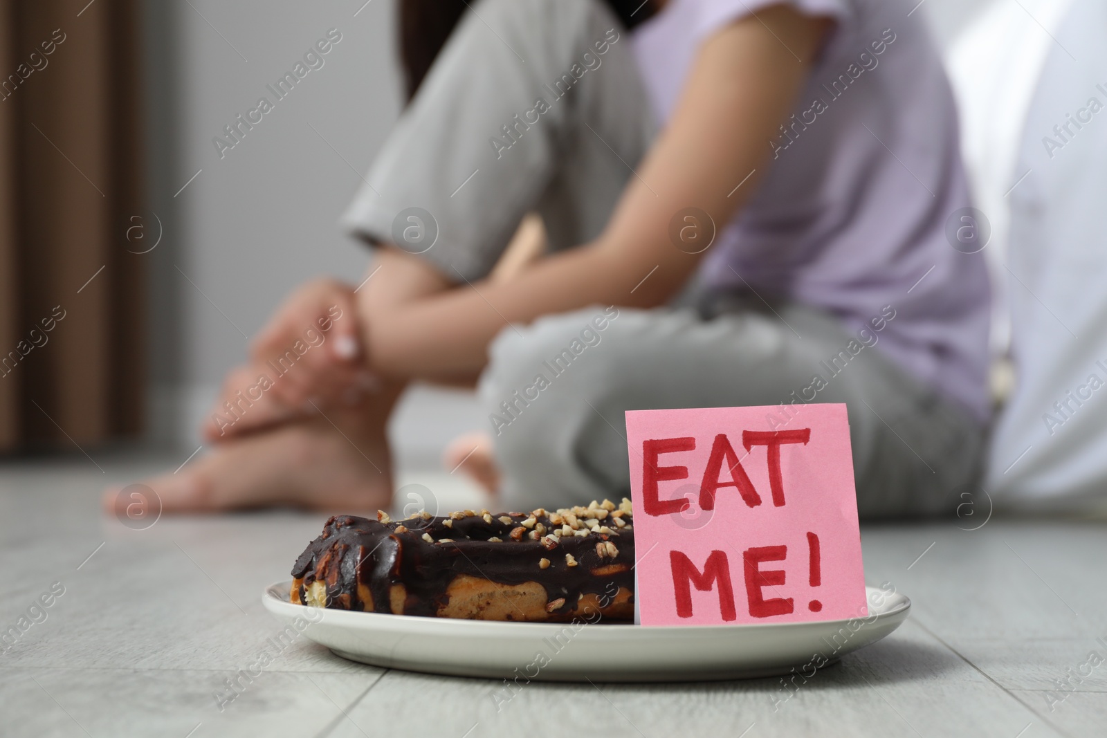 Photo of Eating disorder. Woman sitting near note with words Eat Me and eclair on floor indoors, selective focus