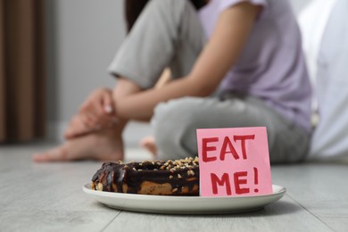Photo of Eating disorder. Woman sitting near note with words Eat Me and eclair on floor indoors, selective focus