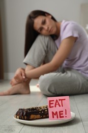 Photo of Eating disorder. Sad woman sitting near note with words Eat Me and eclair on floor indoors, selective focus