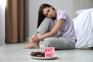 Photo of Eating disorder. Sad woman sitting near note with words Eat Me and eclair on floor indoors, selective focus