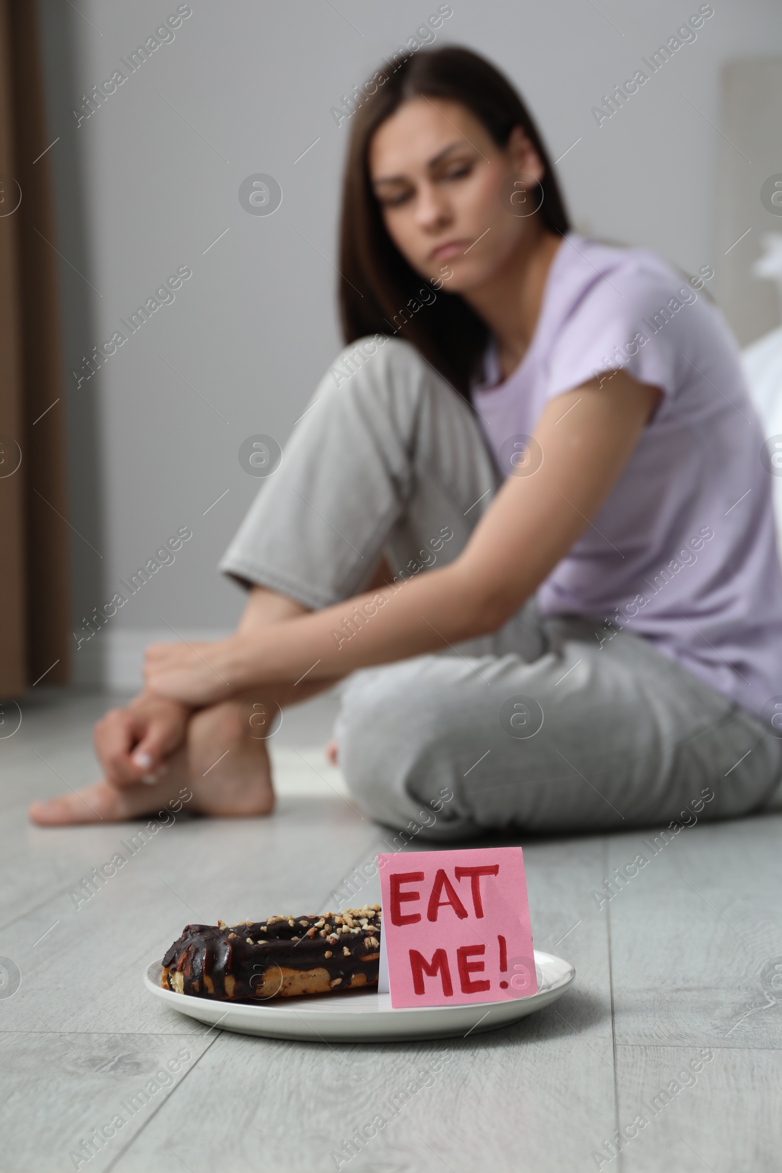 Photo of Eating disorder. Sad woman sitting near note with words Eat Me and eclair on floor indoors, selective focus