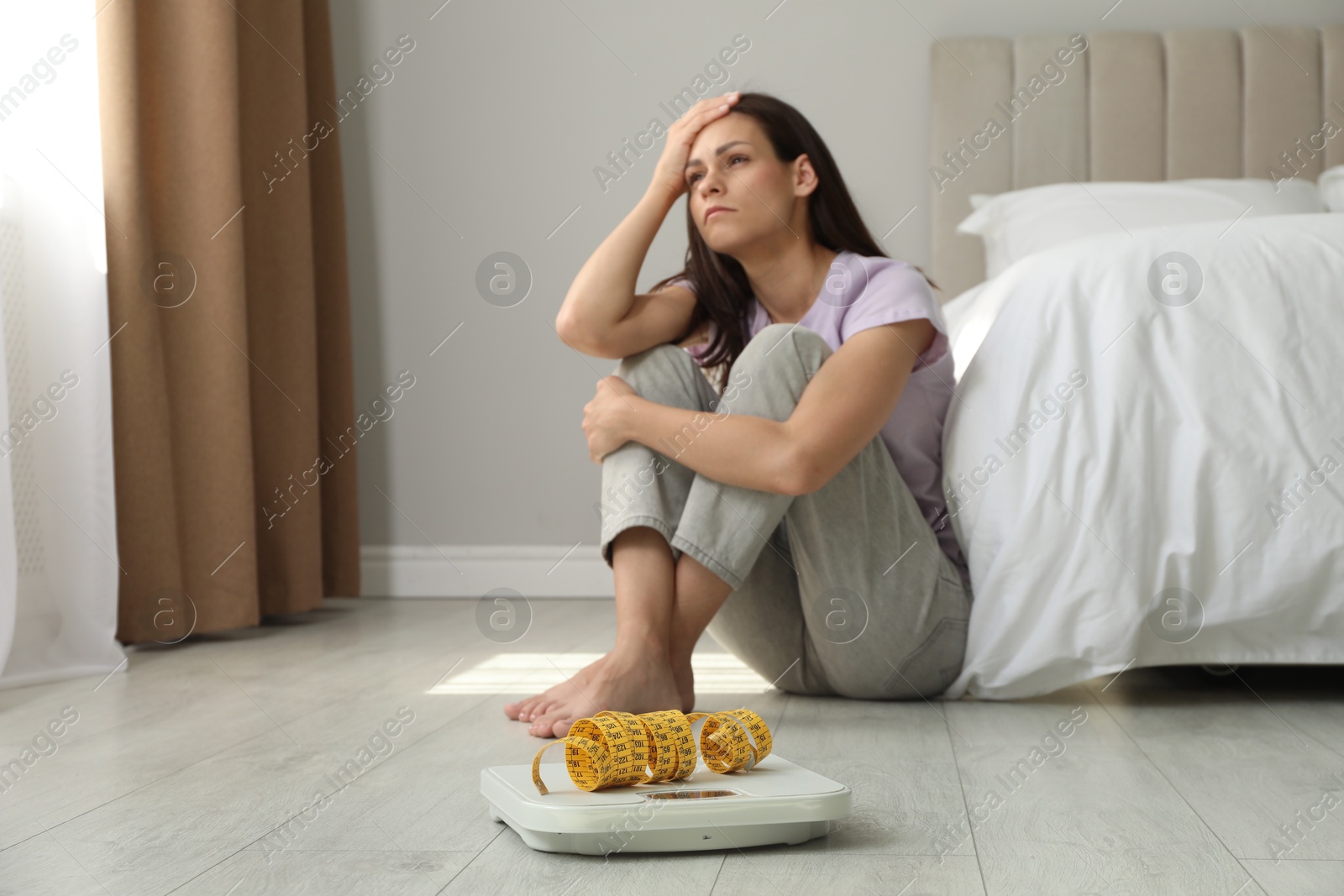 Photo of Eating disorder. Sad woman sitting near scale and measuring tape on floor indoors