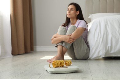 Photo of Eating disorder. Sad woman sitting near scale and measuring tape on floor indoors