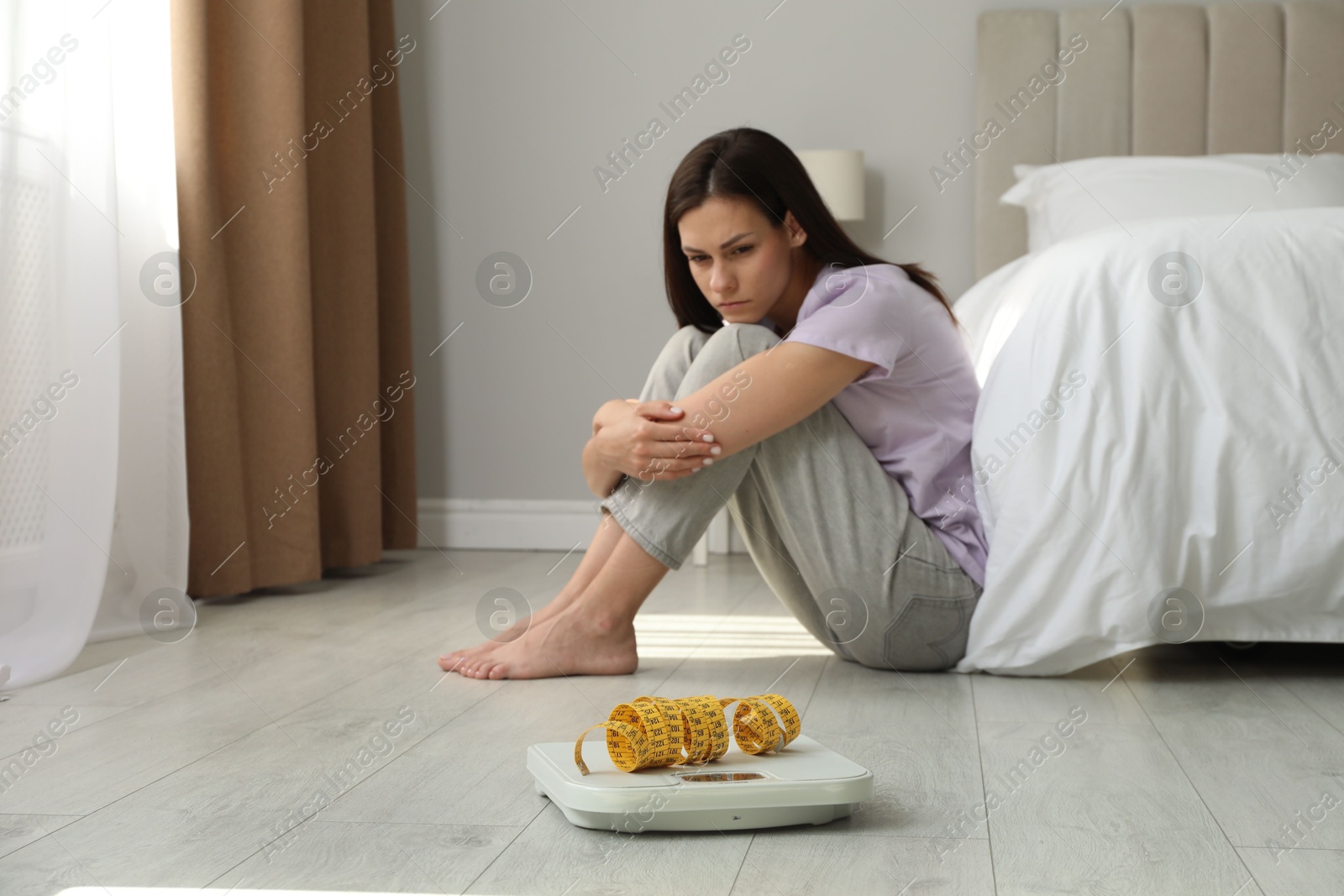 Photo of Eating disorder. Sad woman sitting near scale and measuring tape on floor indoors