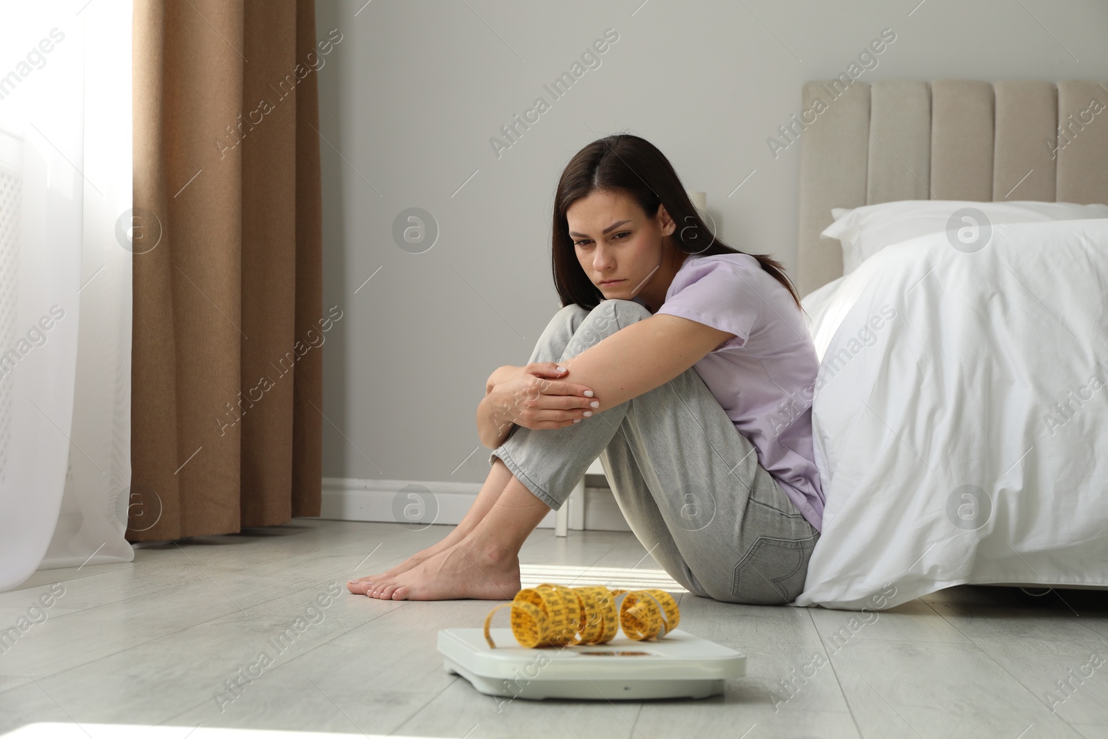 Photo of Eating disorder. Sad woman sitting near scale and measuring tape on floor indoors