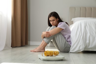 Photo of Eating disorder. Sad woman sitting near scale and measuring tape on floor indoors