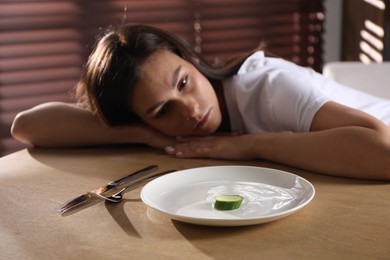 Eating disorder. Sad woman at wooden table with cutlery, cucumber and plate indoors